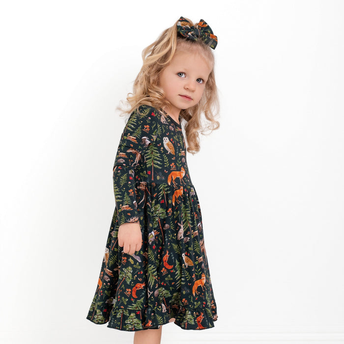 A young child with long blond hair wears the Jammie Session "Wood You Be My Friend" Long Sleeve Big Kid Twirl Dress, featuring a forest animal print and matching bow, standing against a plain white background.