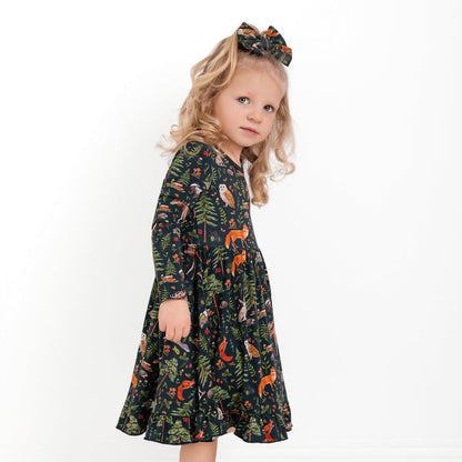 A young child with long blond hair wears the Jammie Session "Wood You Be My Friend" Long Sleeve Big Kid Twirl Dress, featuring a forest animal print and matching bow, standing against a plain white background.