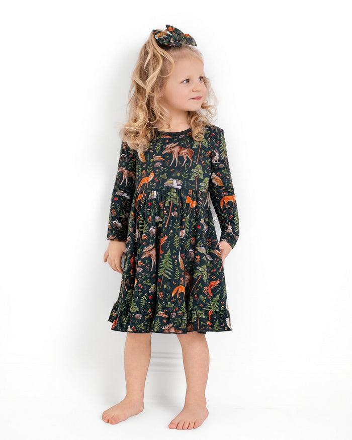 A young girl with blonde curls, barefoot against a white background, wears the Jammie Session "Wood You Be My Friend" long sleeve big kid twirl dress with a forest animal print and a matching hair bow.