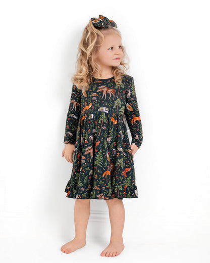A young girl with blonde curls, barefoot against a white background, wears the Jammie Session "Wood You Be My Friend" long sleeve big kid twirl dress with a forest animal print and a matching hair bow.
