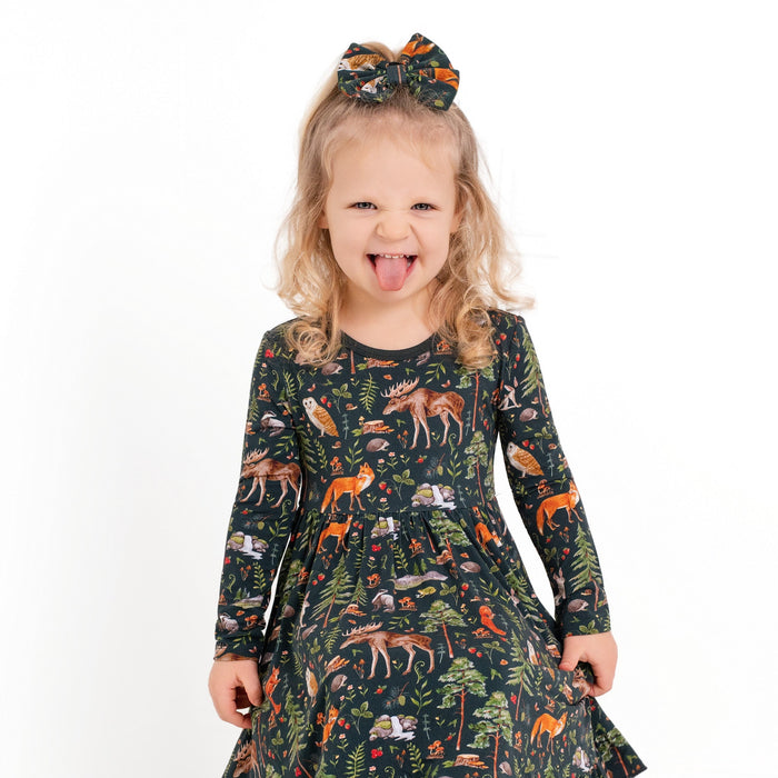 A young girl with light hair in a bow wears the Jammie Session Wood You Be My Friend Long Sleeve Big Kid Twirl Dress, smiling and sticking out her tongue as she stands against a white background.