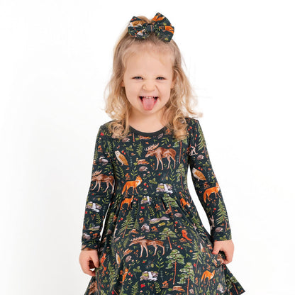 A young girl with light hair in a bow wears the Jammie Session Wood You Be My Friend Long Sleeve Big Kid Twirl Dress, smiling and sticking out her tongue as she stands against a white background.