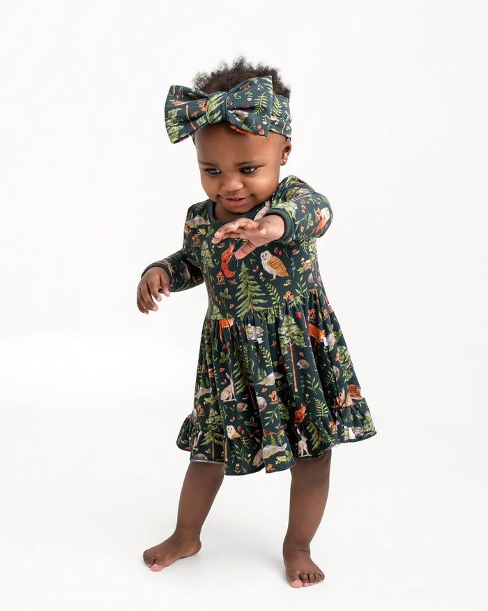 A toddler in a Jammie Session Wood You Be My Friend Long Sleeve Bodysuit Twirl Dress and matching headband stands barefoot against a plain white background, reaching one arm forward.