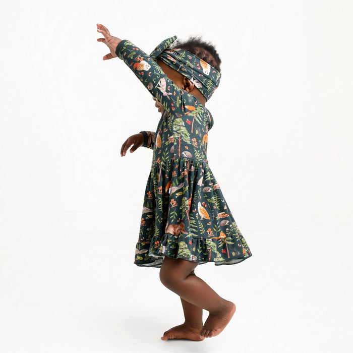 A young child in a Jammie Session "Wood You Be My Friend" Long Sleeve Bodysuit Twirl Dress and matching headband stands barefoot against a plain white background, arms raised and face partially obscured.