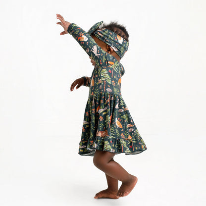 A young child in a Jammie Session "Wood You Be My Friend" Long Sleeve Bodysuit Twirl Dress and matching headband stands barefoot against a plain white background, arms raised and face partially obscured.