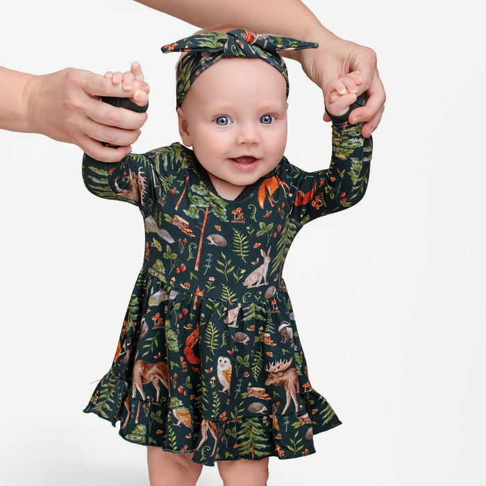 A baby wears the Jammie Session "Wood You Be My Friend" long sleeve bodysuit twirl dress with a forest animal print, plus a matching headband, held by an adult on a plain white background.
