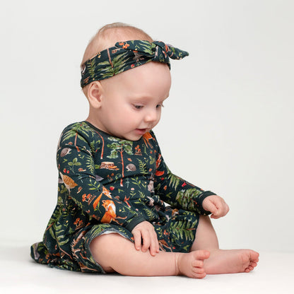 A baby in a Jammie Session Wood You Be My Friend Long Sleeve Bodysuit Twirl Dress with a forest animal print headband sits on the floor against a plain background.