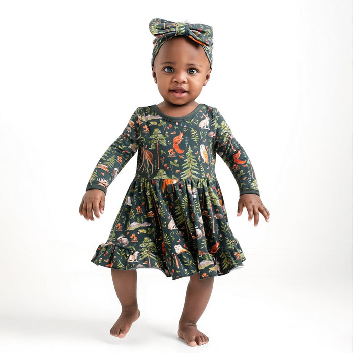 A baby stands barefoot in a Jammie Session "Wood You Be My Friend" Long Sleeve Bodysuit Twirl Dress with matching headband, featuring a forest animal and tree print, against a plain white background.