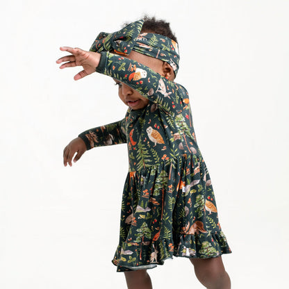 A young child in a Jammie Session Wood You Be My Friend Long Sleeve Bodysuit Twirl Dress with a forest animal print and headband stands sideways, one arm raised, against a plain background.