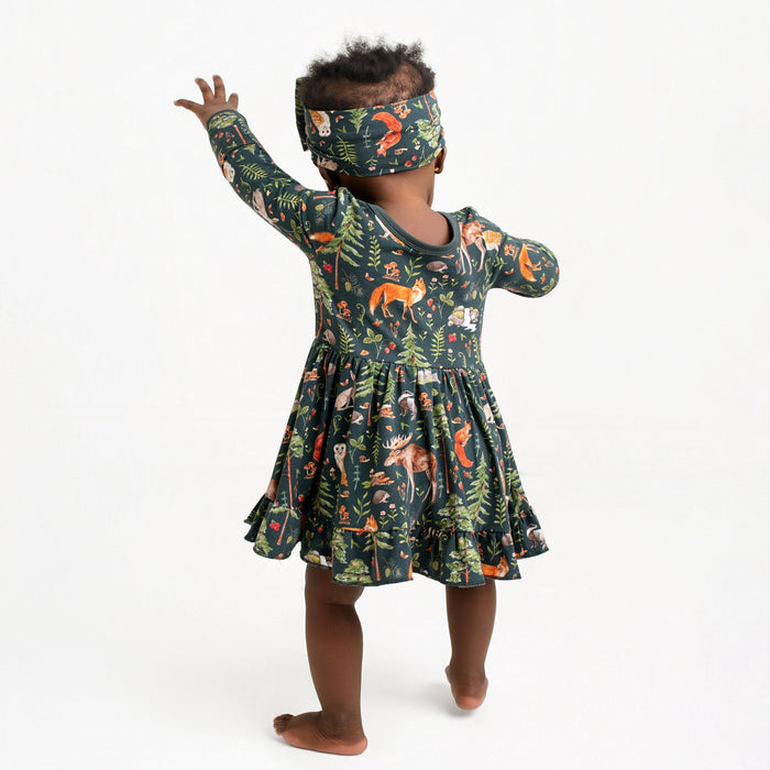A toddler with dark skin and curly hair stands barefoot, facing away, wearing a matching headband and the Jammie Session "Wood You Be My Friend" Long Sleeve Bodysuit Twirl Dress featuring a charming forest animal print.