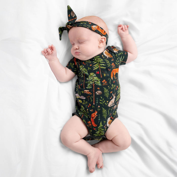 A baby sleeps on a white sheet, wearing Jammie Session‚Äôs Wood You Be My Friend Short Sleeve Bodysuit‚Äîa forest-themed, hypoallergenic bamboo outfit with matching animal and tree print headband.
