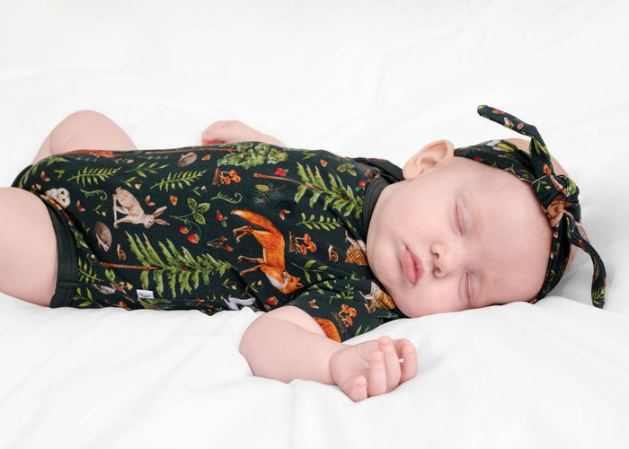 A baby in the Jammie Session "Wood You Be My Friend" short sleeve bodysuit sleeps peacefully on a white bed, wearing a headband with woodland animal prints.
