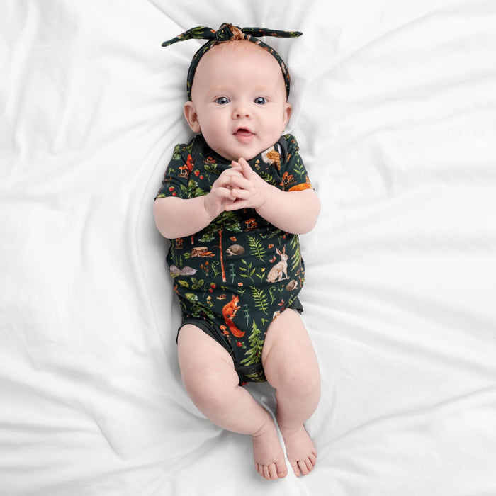 A baby lies on a white bed, wearing the Jammie Session "Wood You Be My Friend" short sleeve bodysuit with animal and plant prints, paired with a matching headband, looking up with hands clasped.
